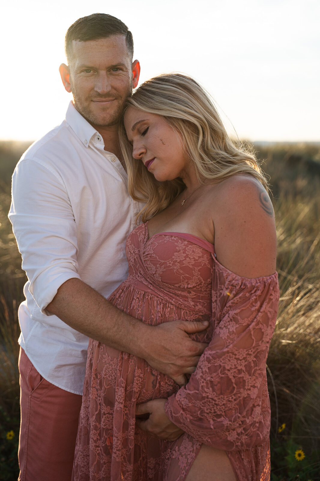 A husband holds his pregnant wife close at golden hour; she rests her head against his shoulder, eyes closed, in a field of sea grass