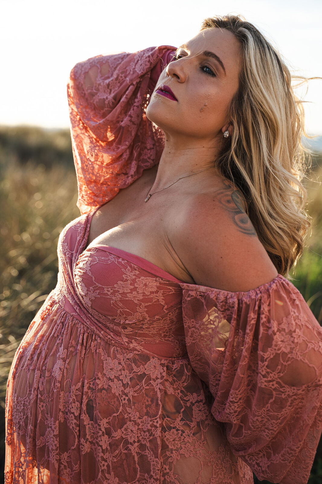 A pregnant woman in profile, hand raised, backlit by golden hour light over coastal grasses
