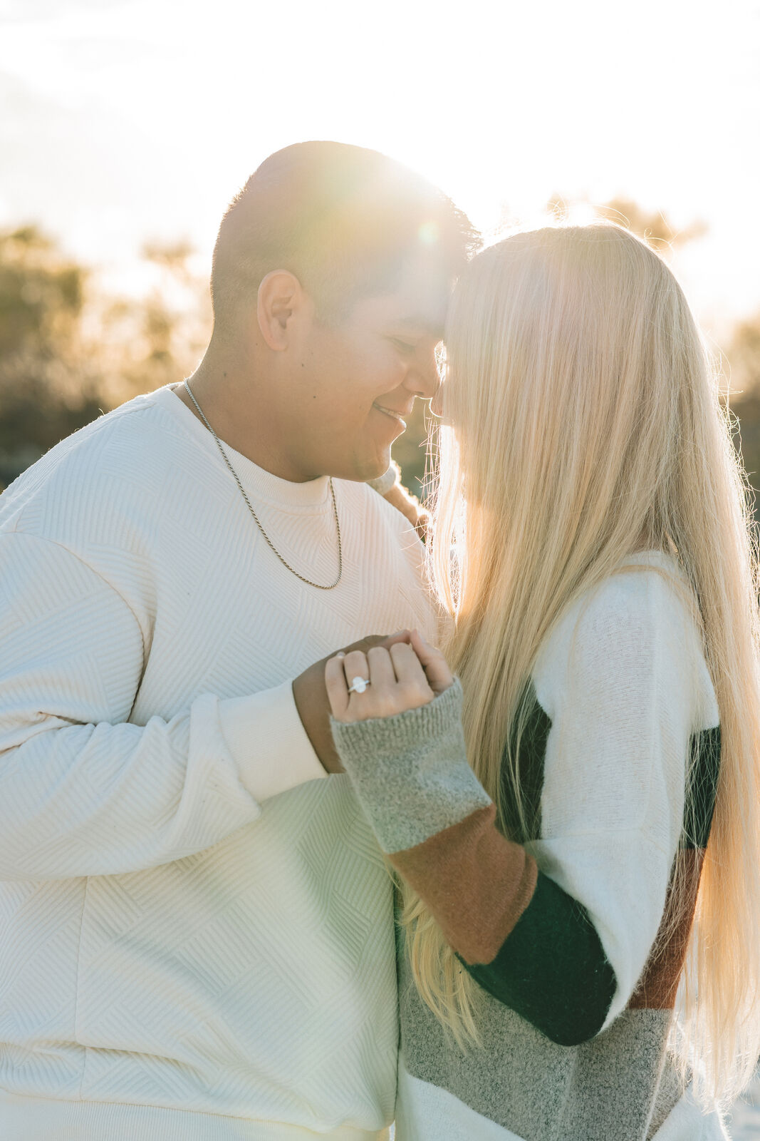 An engaged couple touch foreheads, eyes closed, framed by a sunburst of golden hour light; her engagement ring rests on his shoulder
