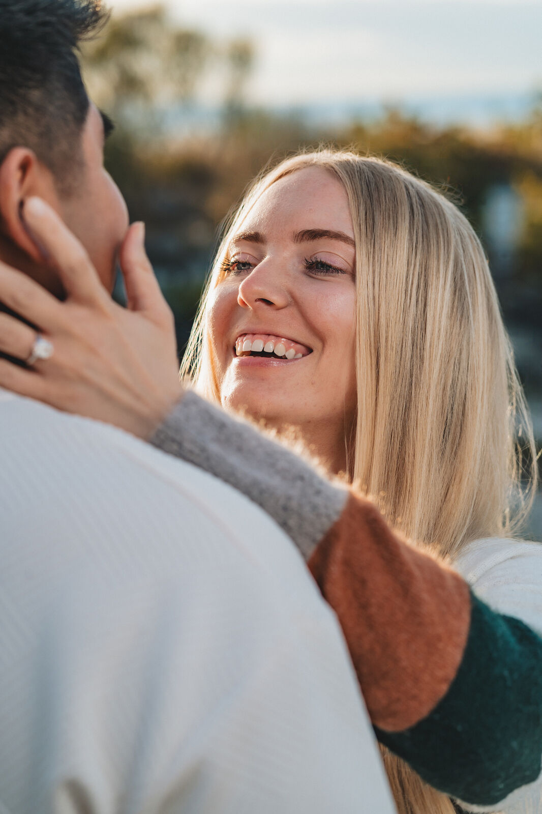 A blonde woman laughs as she looks at her fiancé, her ringed hand resting against his neck, golden hour light behind them