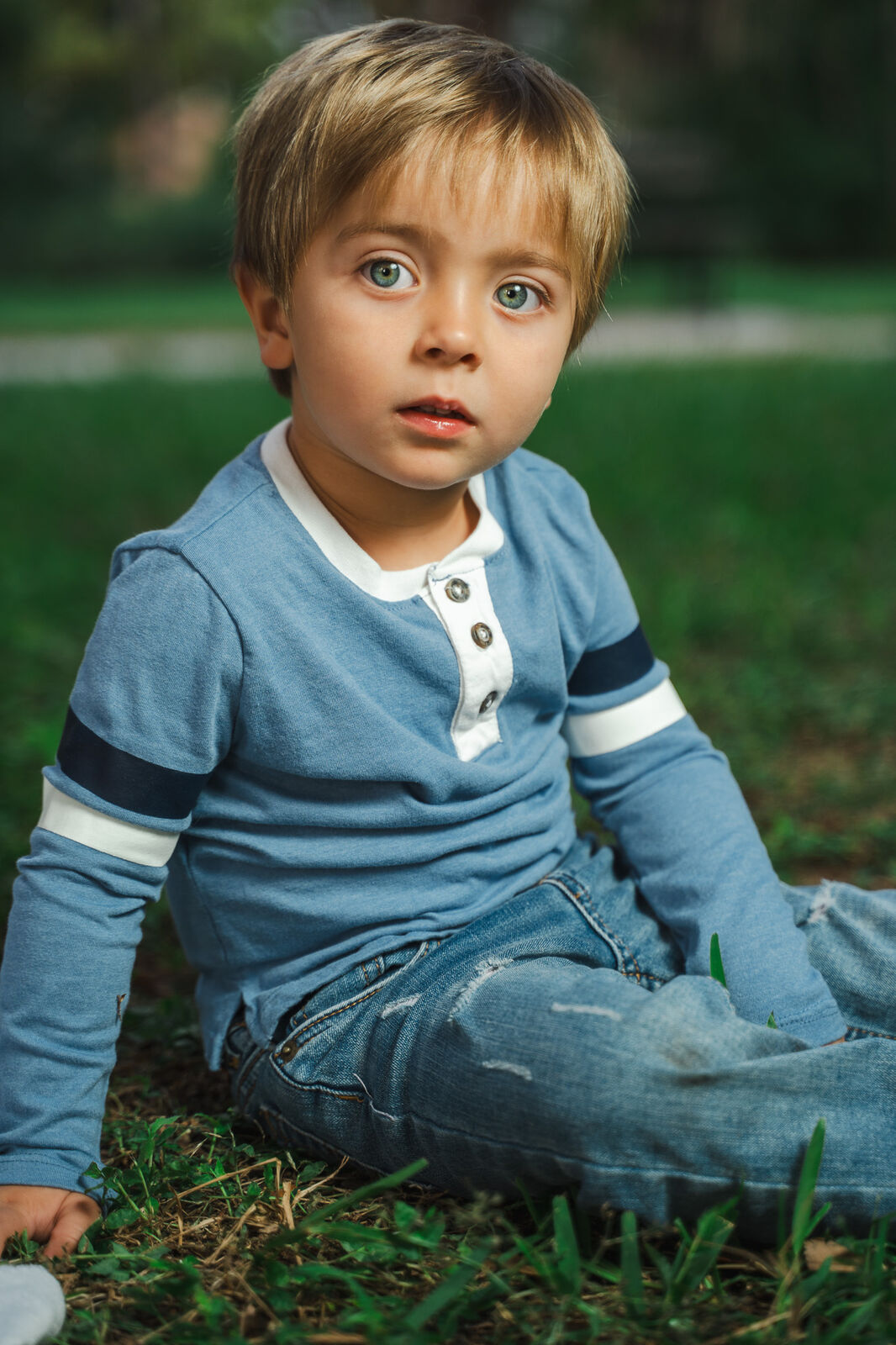 A young boy with bright blue-green eyes sits in the grass at golden hour, wearing a blue henley with white stripes