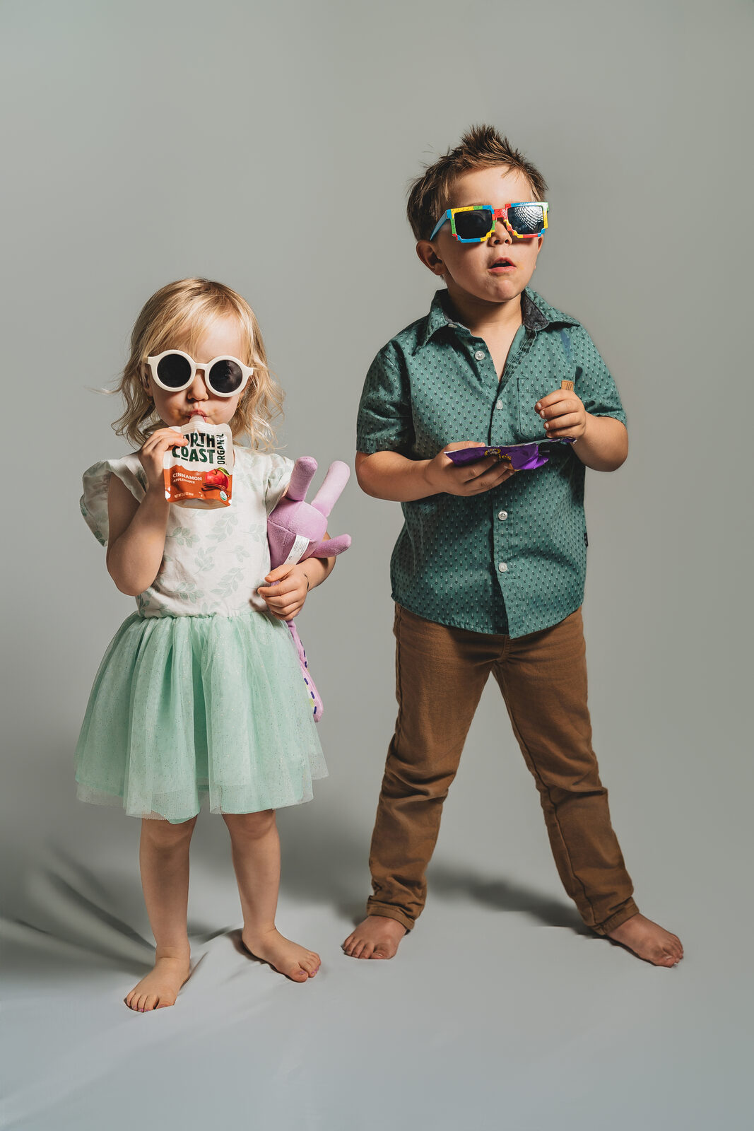 Two young children stand in a studio against a soft gray sweep, both wearing oversized sunglasses, casually snacking — a portrait with personality