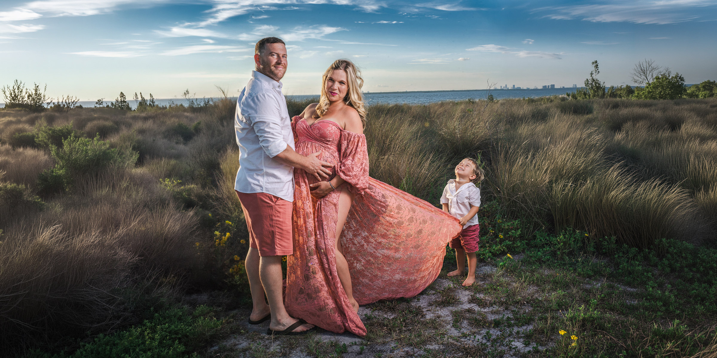 A family on the Tampa Bay dunes at golden hour: a pregnant mother in a flowing pink lace gown stands with her husband; their young son holds the train of her dress, laughing