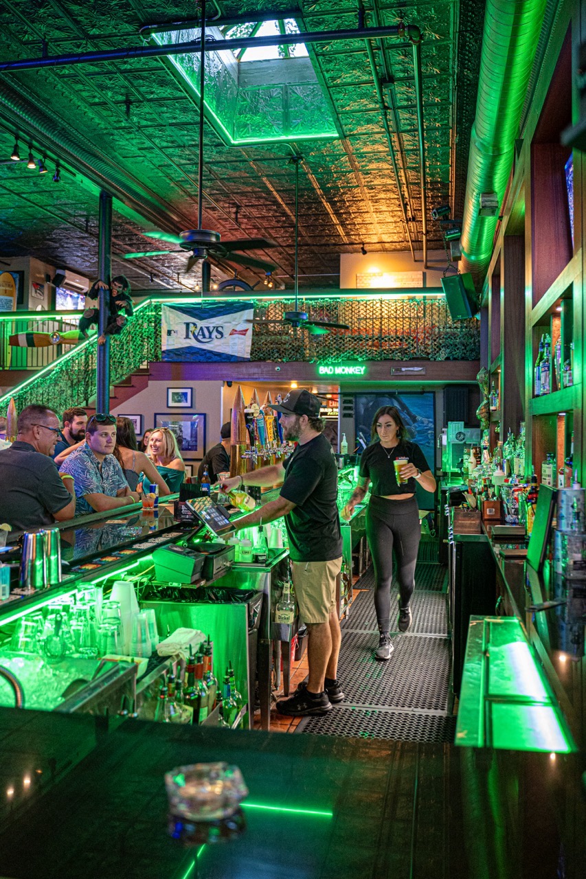Bad Monkey bar interior at full service — bartenders pouring beer, patrons at the bar under green ceiling lights