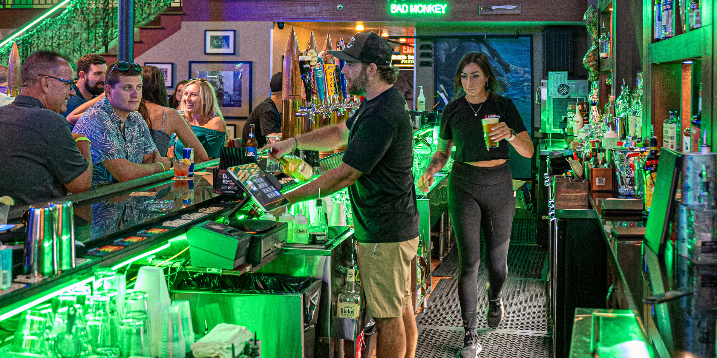 Bad Monkey bar interior at full service — bartenders pouring beer, patrons at the bar under green ceiling lights, Ybor City